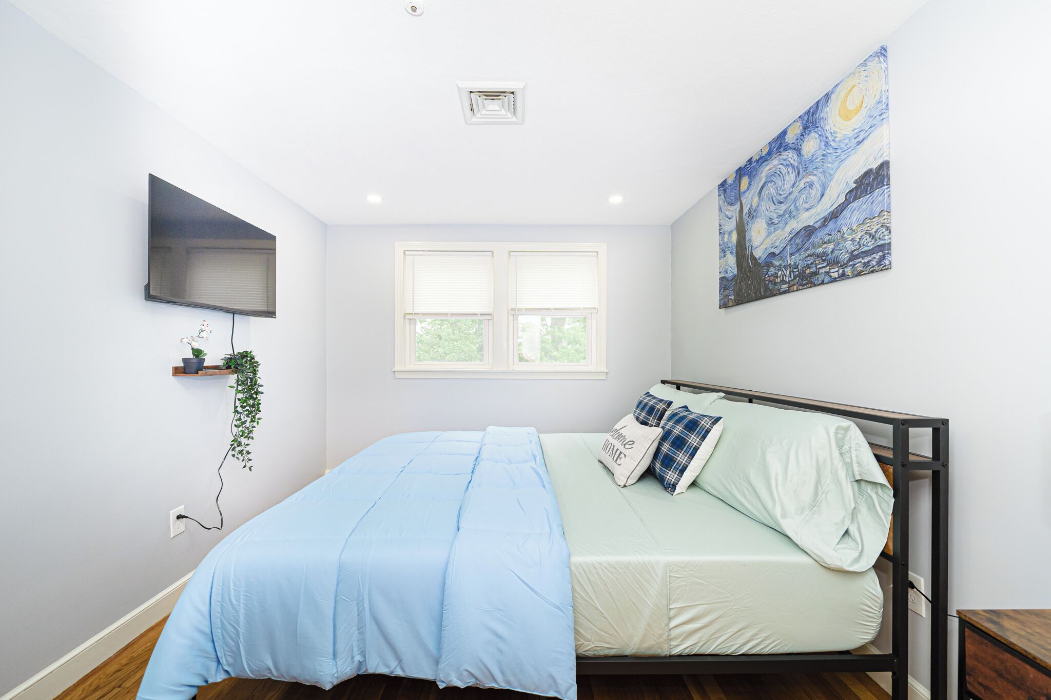 Une chambre à coucher avec un lit, un téléviseur fixé au mur, une fenêtre avec des stores et un tableau au-dessus du lit.