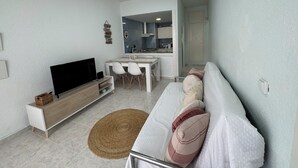 Un salon moderne avec un téléviseur à écran plat, un canapé avec des coussins, une table à manger avec des chaises et, au fond, un coin cuisine.