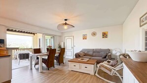Un grand espace de vie comprenant une table à manger, un canapé et un fauteuil à bascule. On y trouve également un ventilateur de plafond et une fenêtre qui laisse entrer la lumière naturelle.