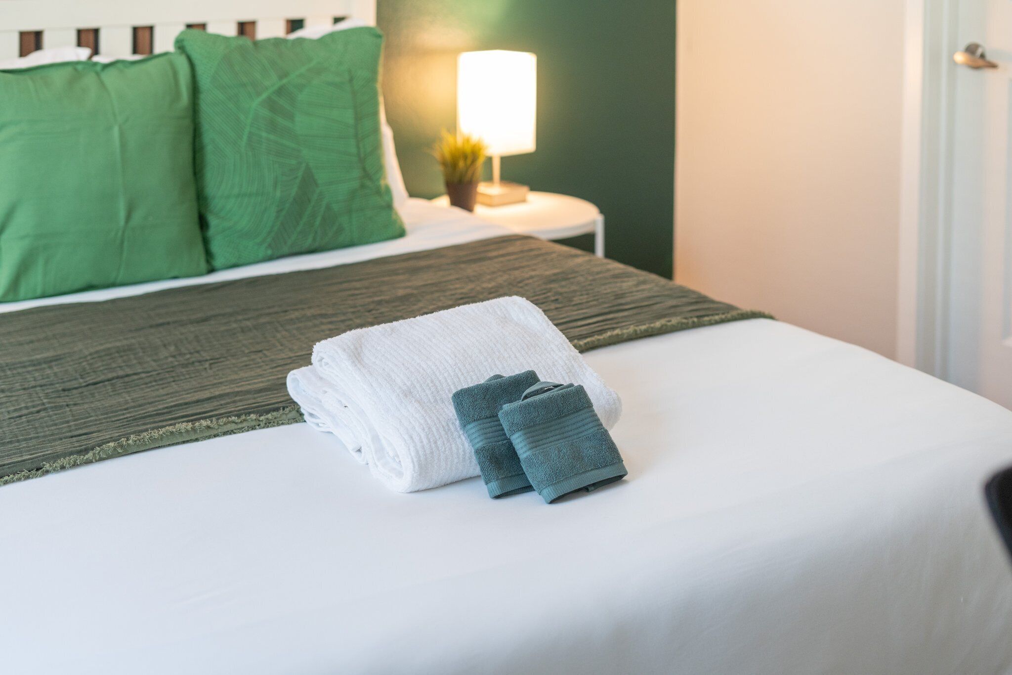 A neatly made bed with a white and green blanket, a folded towel, and a lamp on a bedside table.