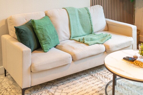 Un canapé beige agrémenté d’un plaid vert et de coussins, une table basse en marbre avec des télécommandes, et un tapis à motifs.