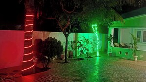 Un cortile con pavimento piastrellato, un vialetto illuminato di verde e un edificio bianco con finestre e una porta.