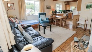 Ein gemütliches Wohnzimmer mit einer schwarzen Ledersofa, einem blauen Sessel und einem Esstisch mit Stühlen. Im Hintergrund ist eine Küche mit Holzschränken und einer Pendelleuchte zu sehen.