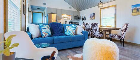 A cozy living room with a blue sofa, white armchair, and a fluffy ottoman. A dining area with patterned chairs is visible in the background.