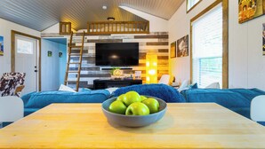 A cozy living room with a wooden table, a bowl of green apples, a TV, and blue sofas. - Shady Grove Tranquil Hill Country Cabin for 8 Near Pedernales Falls (Johnson City)