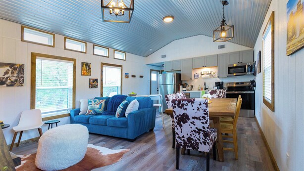 A spacious living area with a blue sofa, a dining table with a cowhide chair, and a kitchen in the background. - Shady Grove Tranquil Hill Country Cabin for 8 Near Pedernales Falls (Johnson City)