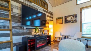 A modern living room with a flat-screen TV, black entertainment unit, white chairs, and a fluffy white ottoman. - Shady Grove Tranquil Hill Country Cabin for 8 Near Pedernales Falls (Johnson City)