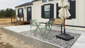 A building with a concrete deck, a wooden bench, two green folding chairs, and a black umbrella. - Shady Grove Tranquil Hill Country Cabin for 8 Near Pedernales Falls (Johnson City)