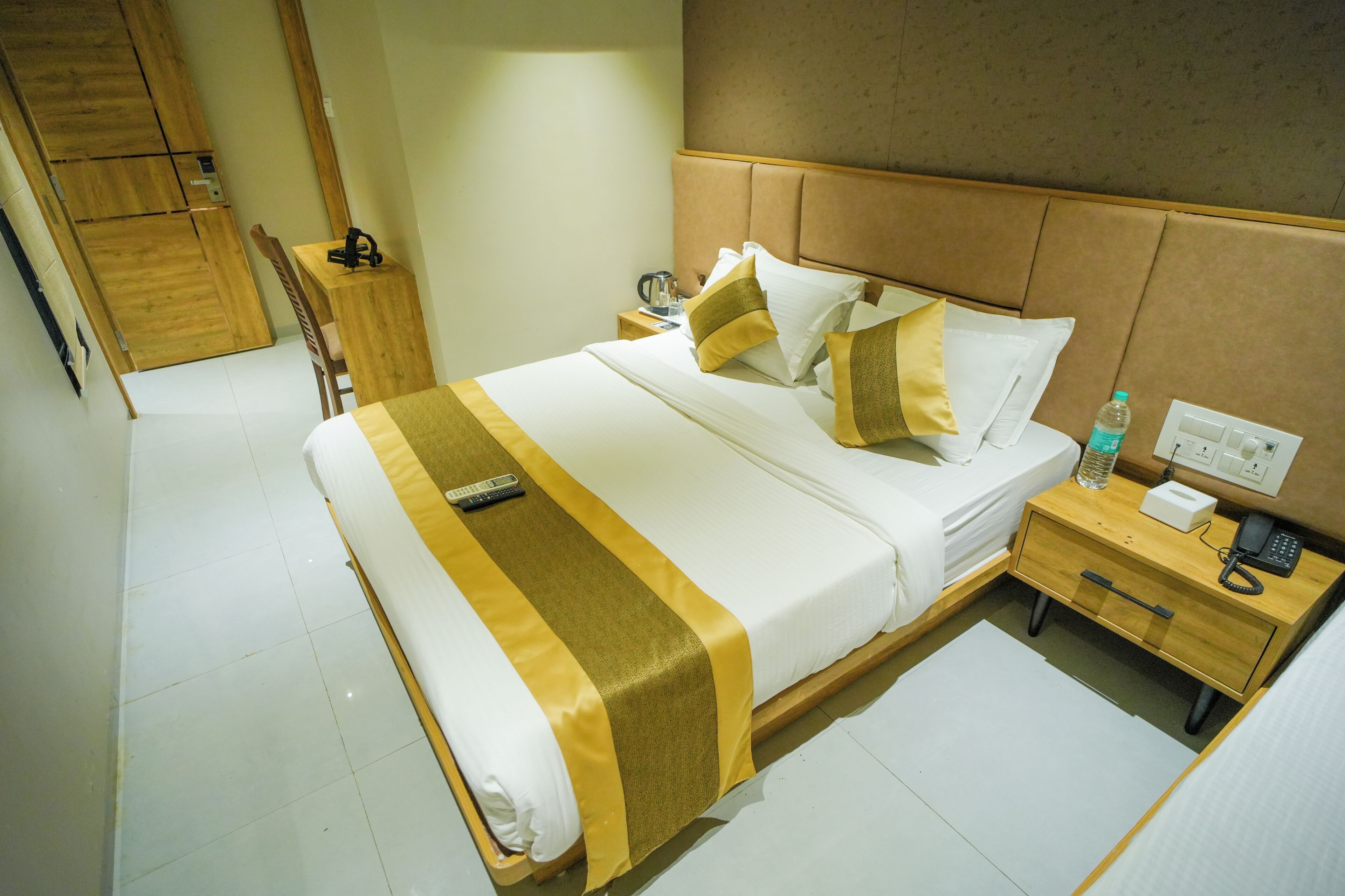 Habitación de hotel con una cama grande, cabecera de madera, mesita de noche con lámpara y un pequeño escritorio con silla.