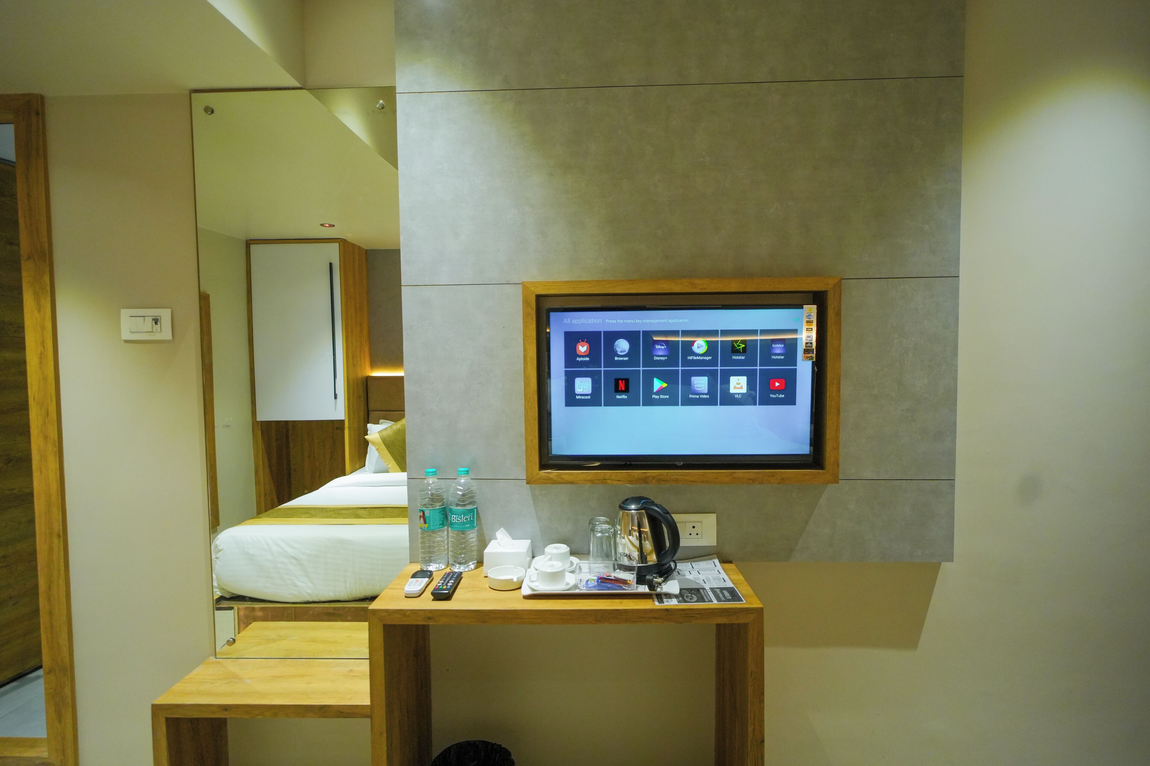 A hotel room with a bed, a bedside table with a remote control, a water bottle, a cup, a kettle, and a mounted television displaying various apps.