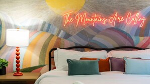 Una habitación de hotel con una cama, almohadas coloridas, un letrero de neón que dice 'The Mountains Are Calling' y una lámpara en una mesita de noche.