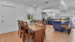 A modern kitchen with white cabinets, a central island with bar seating, and a dining area with wooden chairs and a table. - Charming 3-bedroom house in lovely Naples with AC, Pool and Pet Friendly (Naples)