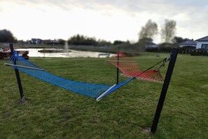 A hammock set up in a grassy area with a pond and a red net in the background. - Holiday home, 3 bedrooms, fishing pond, sauna (Wodnica)