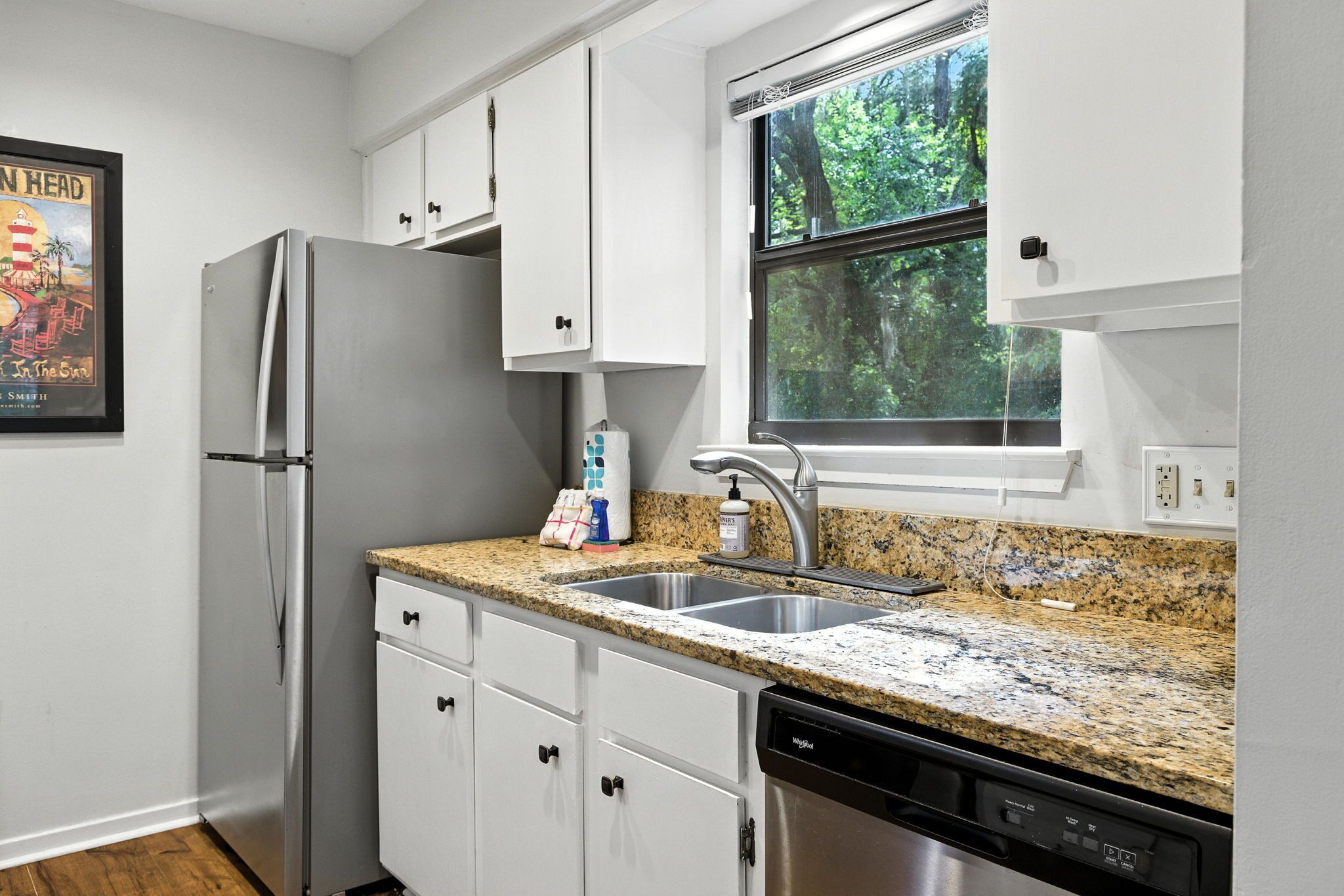 A compact kitchen with white cabinets, a stainless steel refrigerator, a granite countertop, and a window with a view of trees.