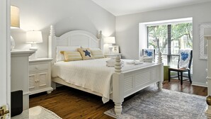 A bedroom with a white bed, two bedside tables with lamps, a window with blinds, and a chair. - Lighthouse Tennis Villas: Walk to both Harbor Town, Shops at Sea Pines STR077622 (Hilton Head)