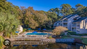 A pool area with a wooden deck, surrounded by trees and a building with balconies. - Lighthouse Tennis Villas: Walk to both Harbor Town, Shops at Sea Pines STR077622 (Hilton Head)