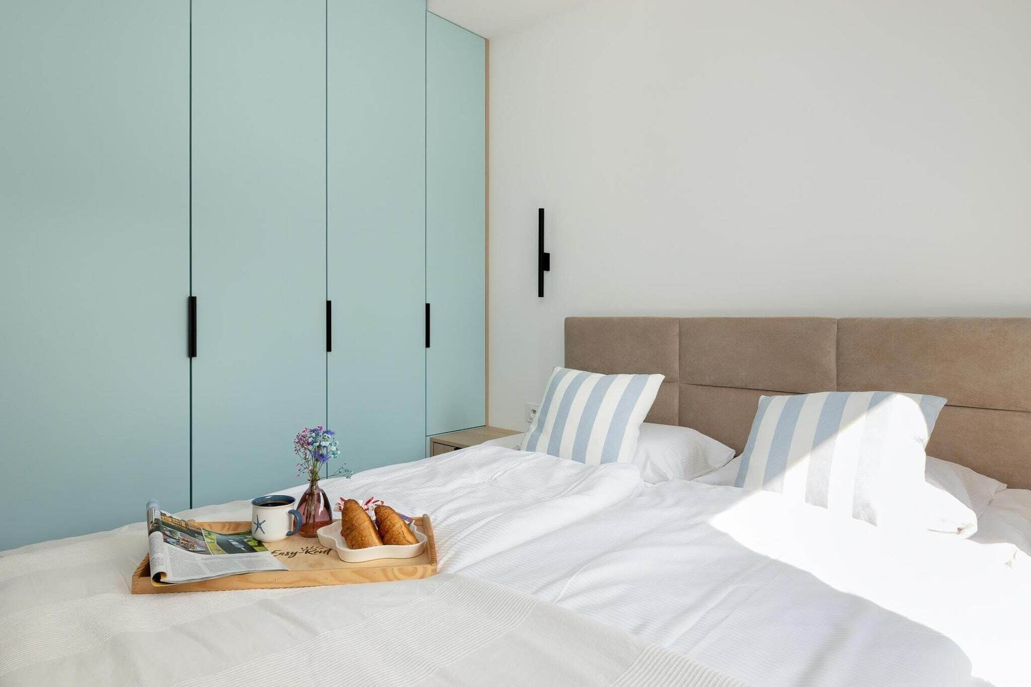 A neatly made bed with white linens, a tray with croissants and a cup of coffee, and a magazine. There is a light blue wardrobe with black handles and a beige headboard with striped pillows.