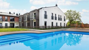 Une piscine avec une terrasse en bois, un bâtiment blanc doté de nombreuses fenêtres et un ensemble de balançoires.