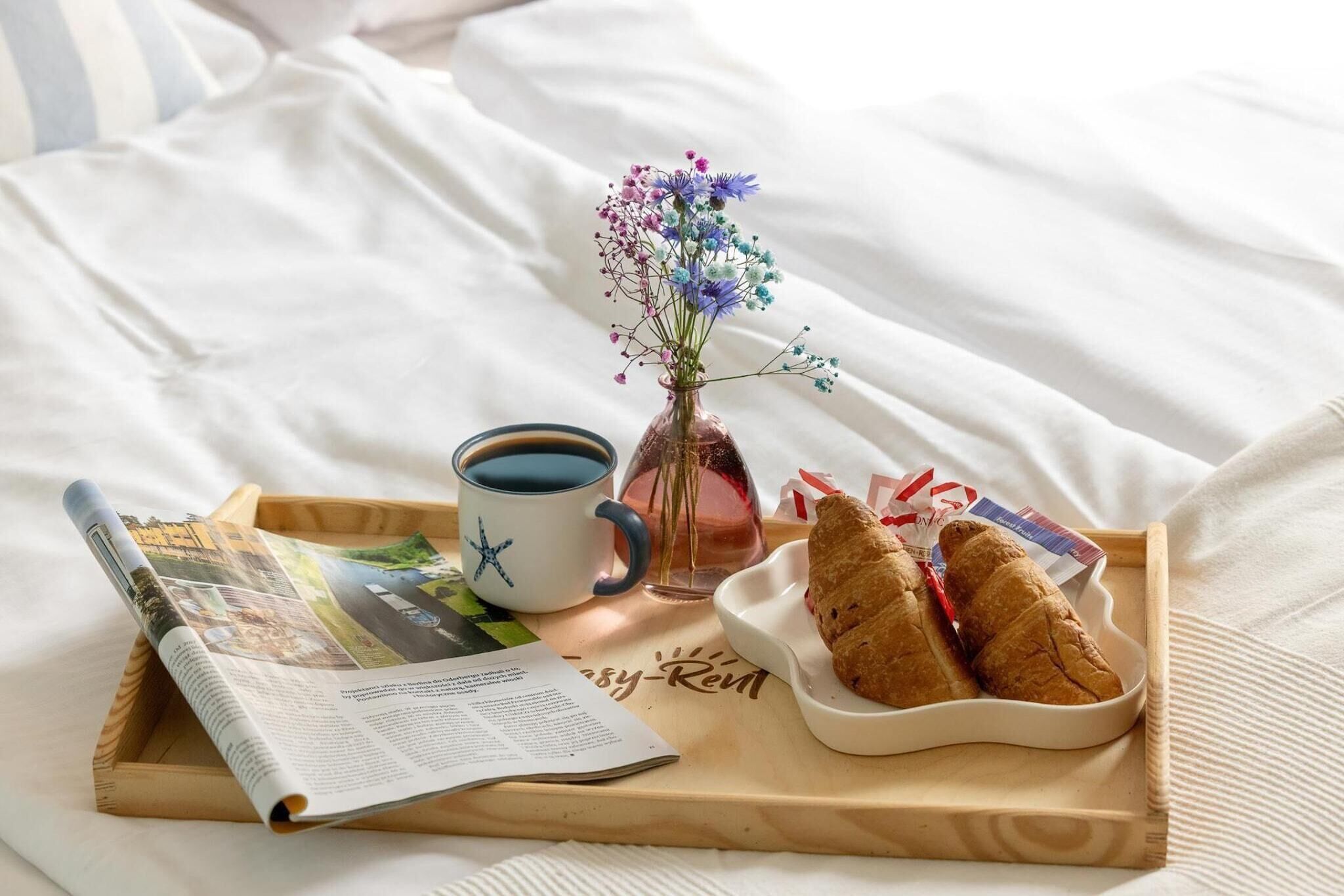 A breakfast tray with bread, a cup of coffee, a vase with flowers, and a magazine on a bed.
