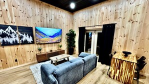 A cozy living room with a wooden paneled wall, a large flat-screen TV, a blue sofa, a wooden coffee table, and a small dining area with a wooden table and chairs.