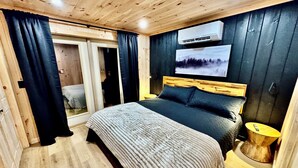 A bedroom with a wooden ceiling, a bed with a dark headboard, a large window with a view of a forest, and an air conditioning unit on the wall.