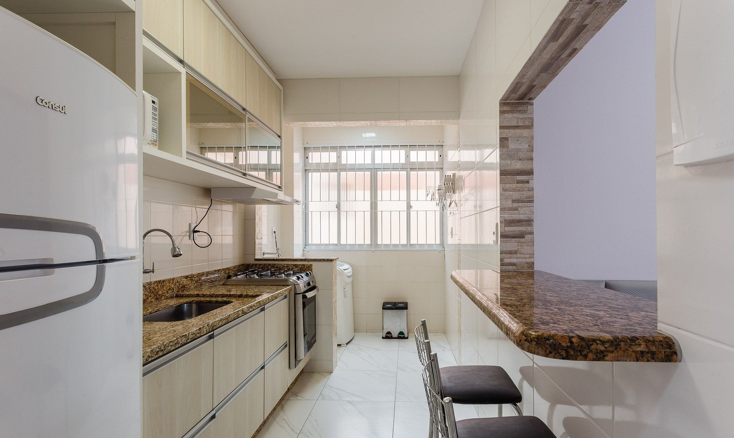 A compact kitchen with a refrigerator, stove, and sink. There is a window with bars and a trash bin.