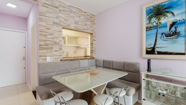 A modern kitchen with a stone backsplash, a dining area with a glass table and cushioned seating, and a framed painting of a tropical scene. - Apt Close to the Beach in Canasvieiras Rfs205 (Florianópolis)