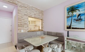 Uma cozinha moderna com painel de backsplash de pedra, uma área de jantar com mesa de vidro e assentos acolchoados, e um quadro com uma cena tropical.