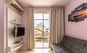 A living room with a flat-screen TV, a sofa, a window with curtains, and a view of buildings outside. - Apt Close to the Beach in Canasvieiras Rfs205 (Florianópolis)