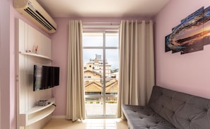Ein Wohnzimmer mit Flachbildfernseher, einer Couch, einem Fenster mit Vorhängen und Blick auf Gebäude.