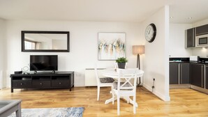 Ein modernes Wohnzimmer mit Flachbildfernseher, Esstisch mit Stühlen, einem Blumentopf und einem Bild an der Wand.