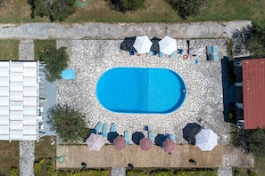 Et luftfoto af en swimmingpool omgivet af liggestole og parasoller, med en stendækket sti, der fører hen til en bygning med rødt tag.