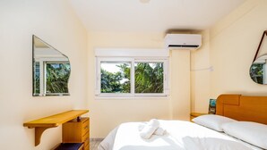 A bedroom with a bed, a wooden desk, a chair, a window with a view of greenery, and an air conditioning unit. - Apart W/barbecue 10min From Jurere Beach Rf203 (Florianópolis)