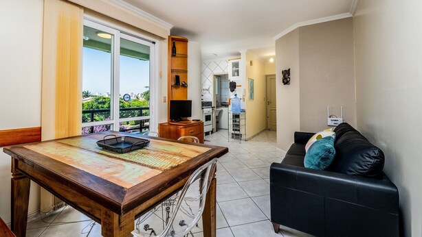 A modern living room with a dining table, a black sofa, and a kitchen area visible through an open door. - Apart W/barbecue 10min From Jurere Beach Rf203 (Florianópolis)