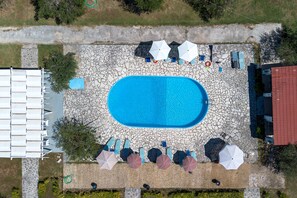 Une vue aérienne d’une piscine entourée de chaises longues et de parasols, avec un chemin de pierre menant à un bâtiment dont le toit est rouge.