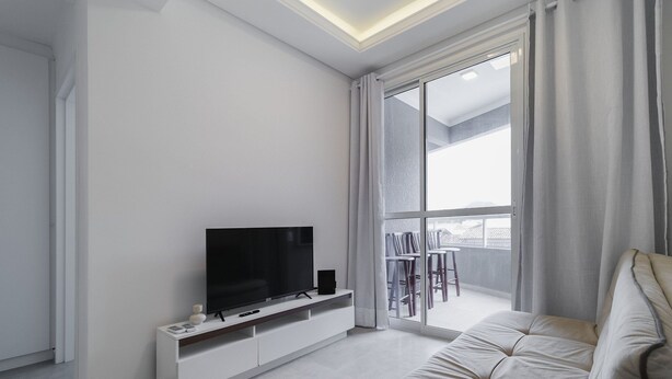 A modern living room with a flat-screen TV on a white console, a large window with curtains, and a balcony with outdoor furniture. - Apt w/ BBQ 300m From the Beach in Floripa Res201 (Florianópolis)