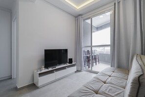 Una sala de estar moderna con un televisor de pantalla plana sobre un mueble blanco, un ventanal con cortinas y un balcón con mobiliario exterior.