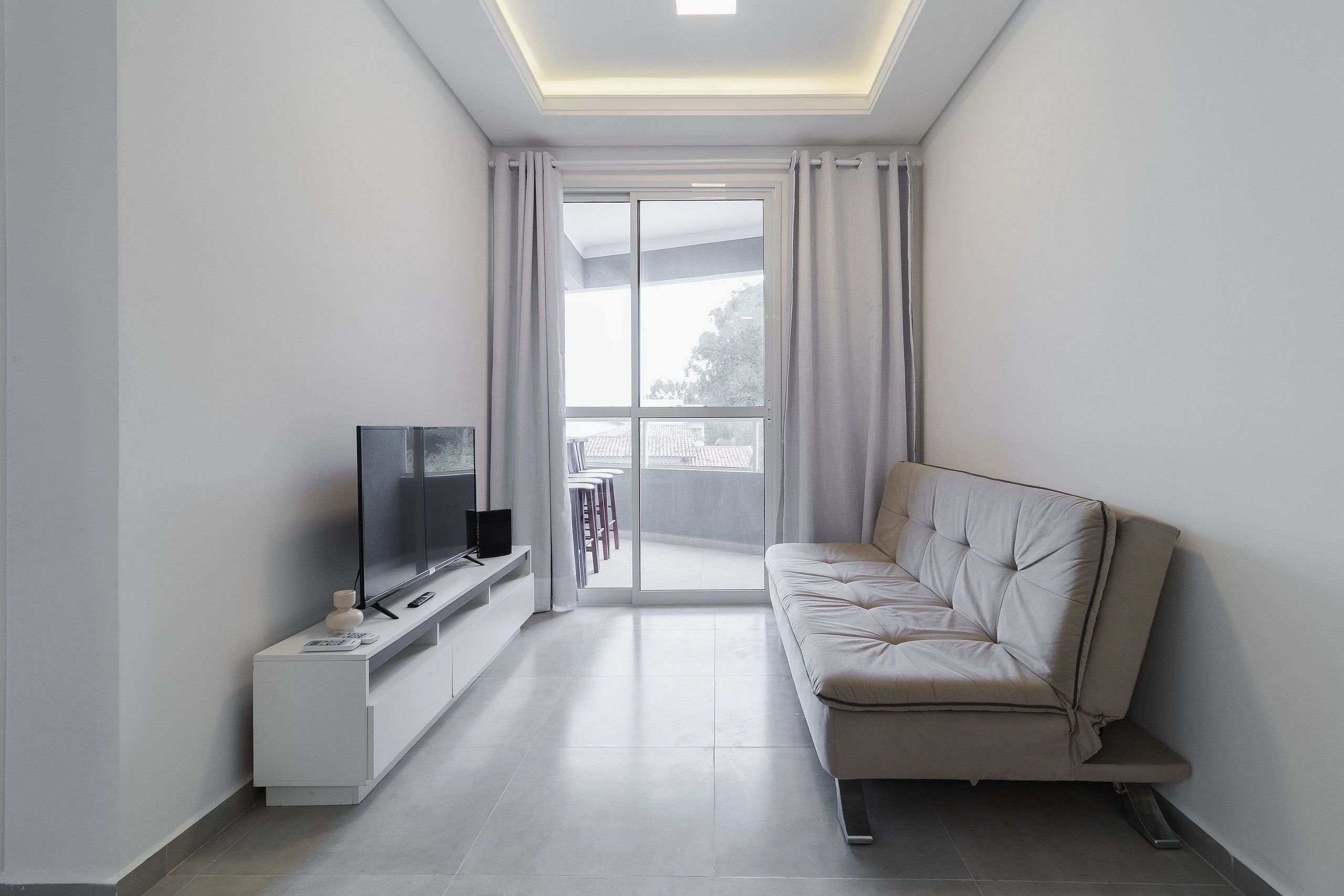 Una sala de estar moderna con un televisor de pantalla plana sobre un mueble blanco, un sofá color beige y un ventanal corredizo que da a un balcón.