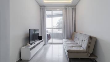 Una sala de estar moderna con un televisor de pantalla plana sobre un mueble blanco, un sofá color beige y un ventanal corredizo que da a un balcón.