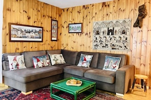 A living room with a sectional sofa, a green coffee table, and wooden panel walls with framed pictures.