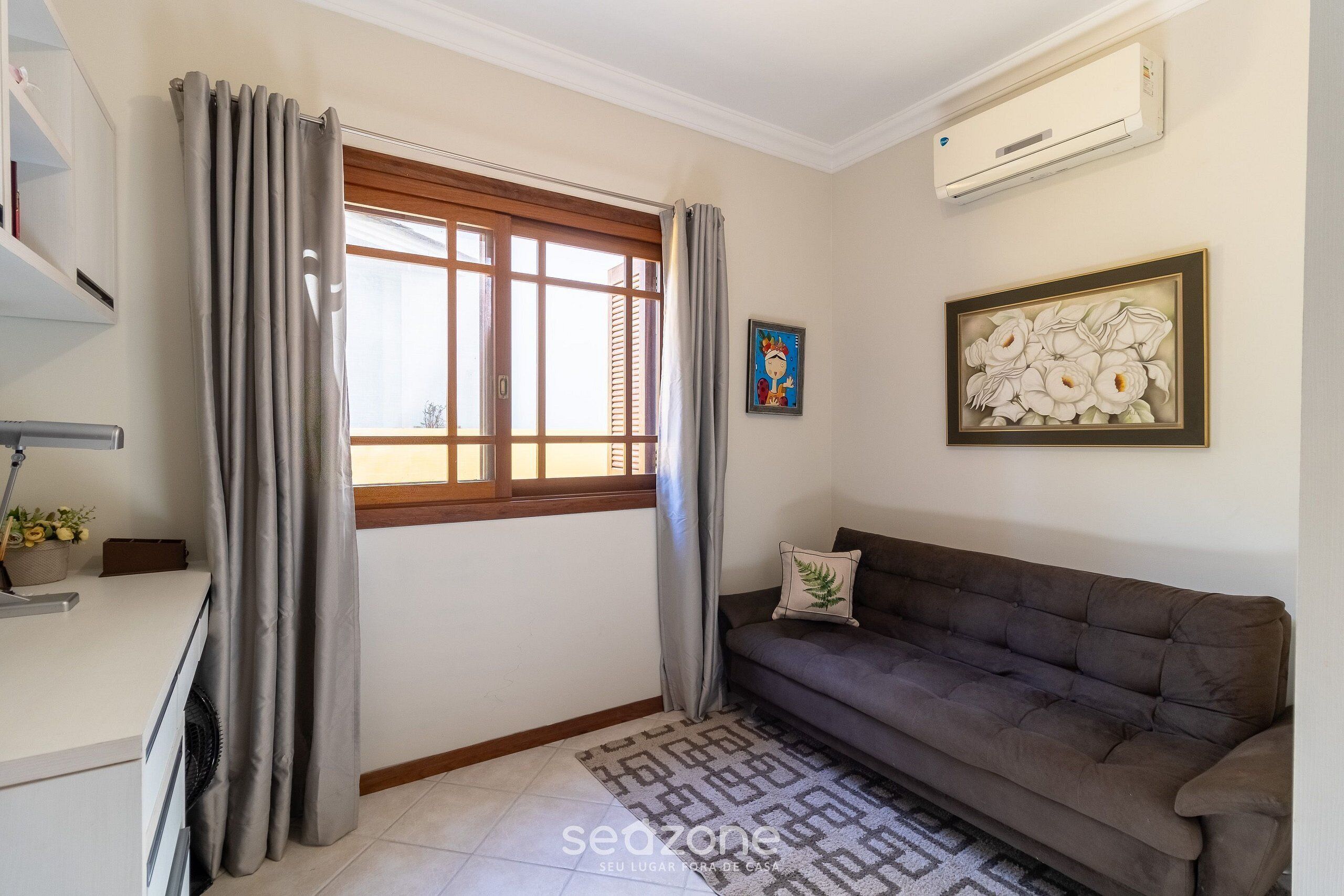 Una sala de estar con sofá, alfombra estampada, ventana con cortinas, aire acondicionado de pared y una obra de arte enmarcada.