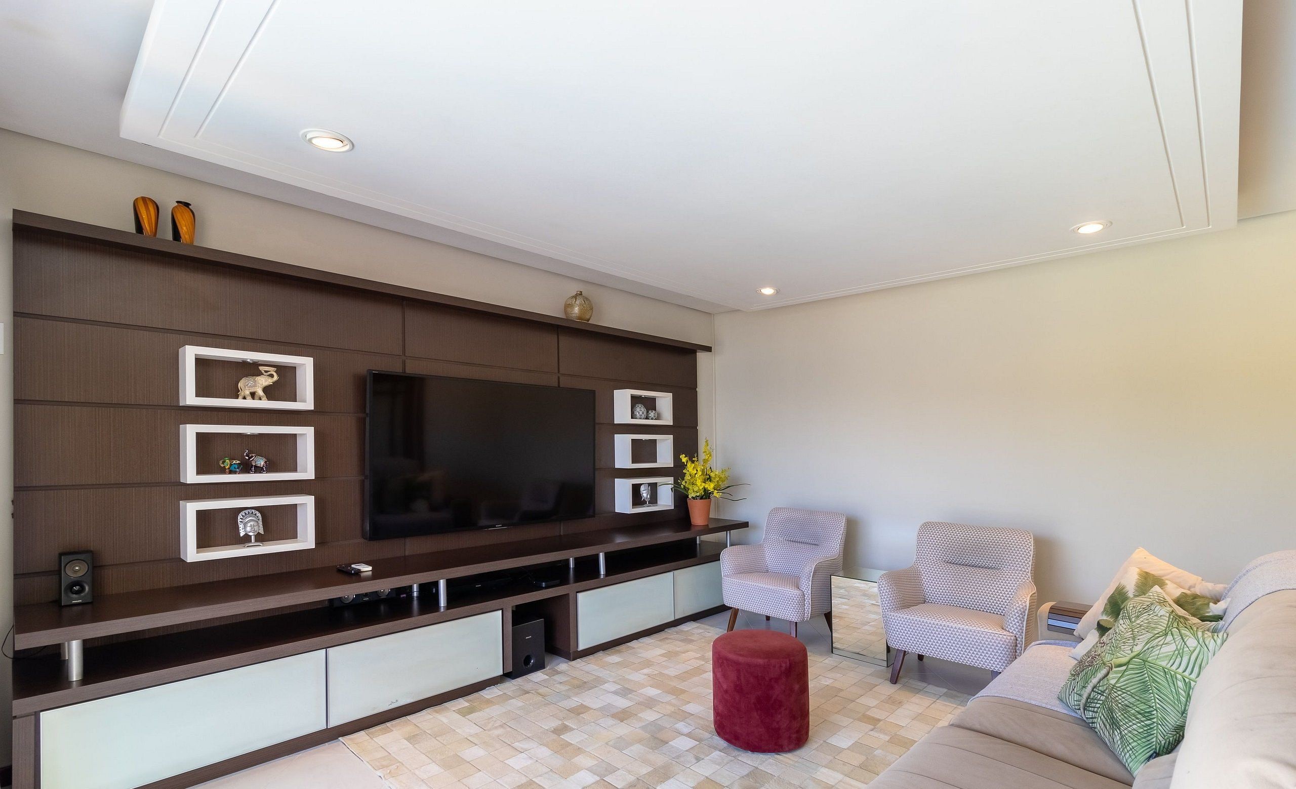 A modern living room with a large flat-screen TV, built-in shelving, and a cozy seating area with a red ottoman and patterned chairs.