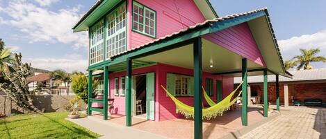 A two-story house with a pink and green exterior, a covered porch with a hammock, and a brick pathway leading to the entrance.