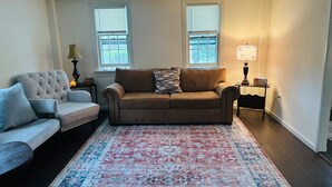 A living room with a beige sofa, a light grey armchair, a patterned rug, two windows with blinds, and a small table with a lamp and a tissue box.