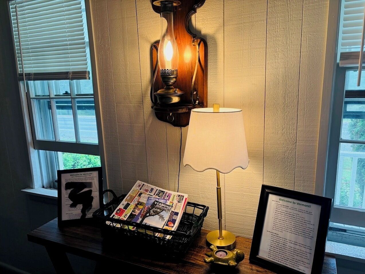 Ein Zimmer mit einer Wandlampe, einem Tisch mit Lampe, einem Magazin-Korb und einem Willkommensschild.