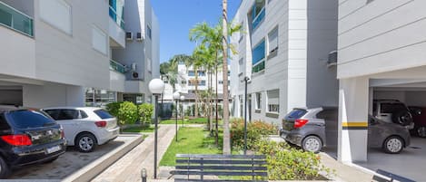 A modern urban courtyard with a paved walkway, benches, parked cars, and palm trees.