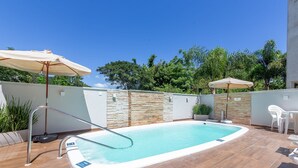 A swimming pool with a metal ladder, surrounded by tiled flooring and stone walls. There are two umbrellas and a white chair visible. - Moderno Apto c/ Piscina em Canasvieiras RCP202 (Florianópolis)