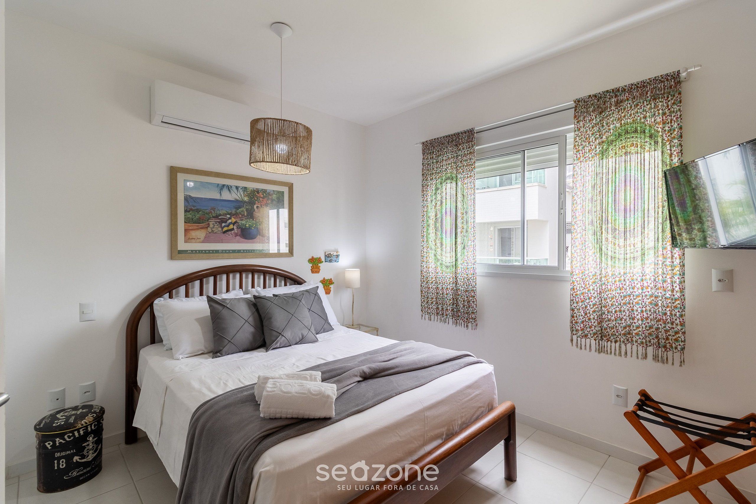 A bedroom with a bed, a hanging light fixture, a window with curtains, a wall-mounted TV, and a small trash bin.