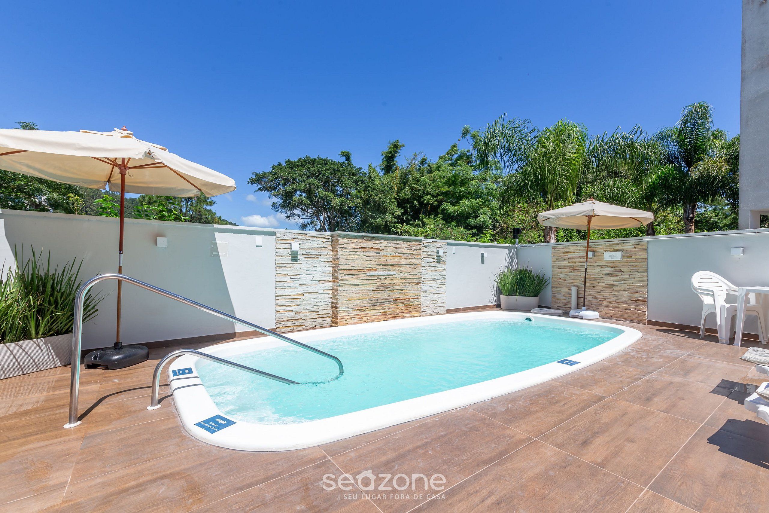 Piscina con una escalera de metal, rodeada de suelo de baldosas y paredes de piedra. Se ven dos sombrillas y una silla blanca.