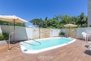 A swimming pool with a metal ladder, surrounded by tiled flooring and stone walls. There are two umbrellas and a white chair visible. - Moderno Apto c/ Piscina em Canasvieiras RCP202 (Florianópolis)